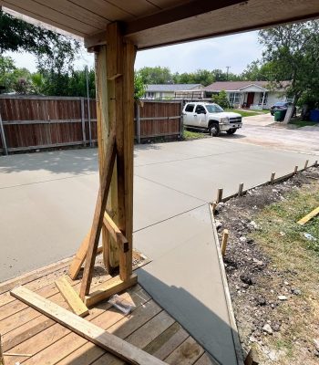 Freshly poured concrete driveway beside a house with a wooden porch, suburban street with parked cars and trees.